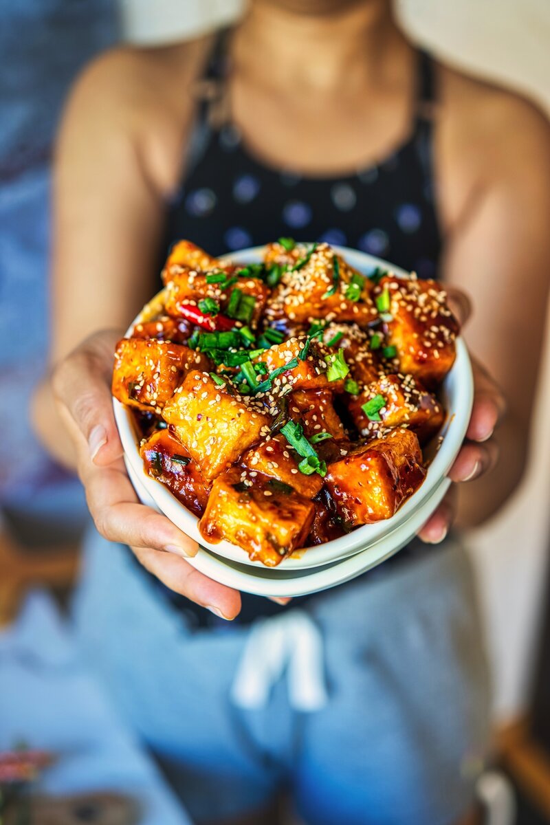 Hands holding a bowl of crispy soy garlic tofu with sesame seeds and scallions, close-up food portrait