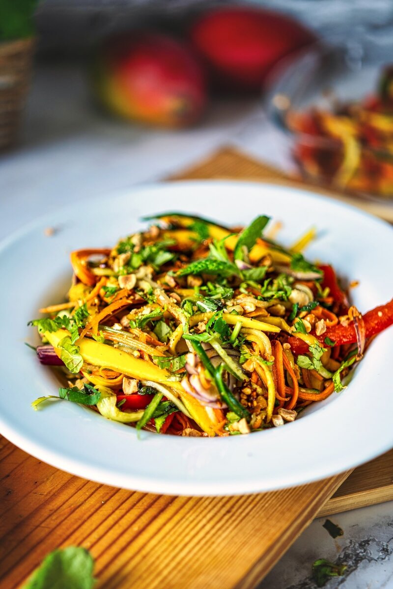Plate of mango salad with julienned vegetables, herbs, and nuts, served on a white plate on a wooden board.
