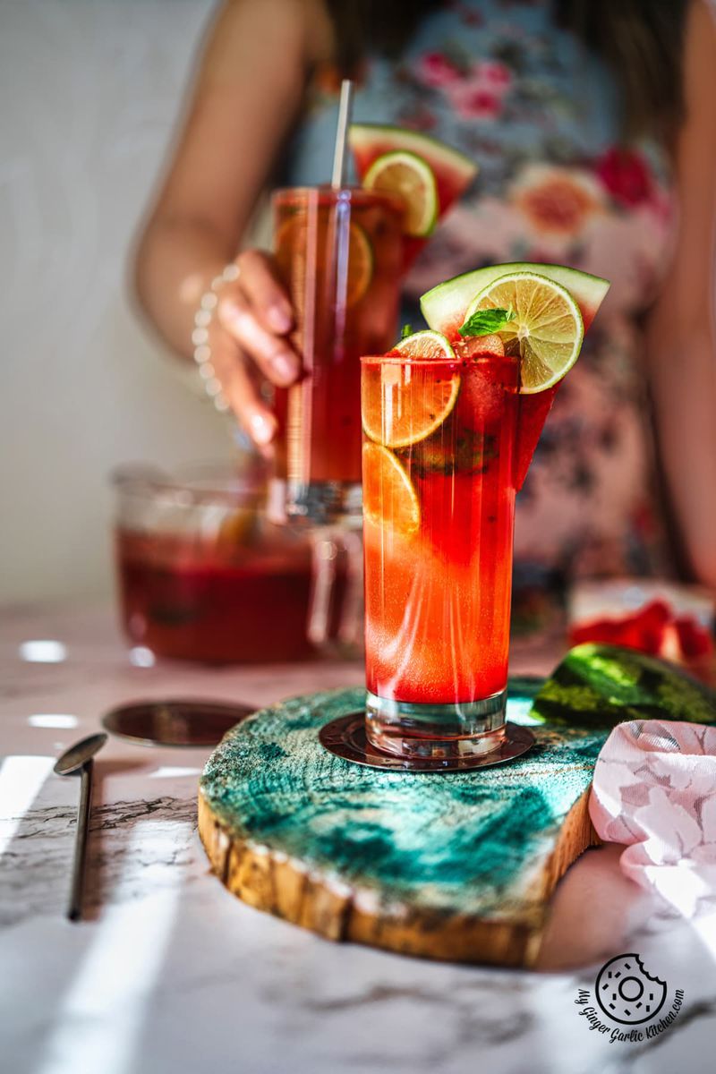 watermelon mojito glass decoarated with lime and watermelon slice and a female holding a glass in the background